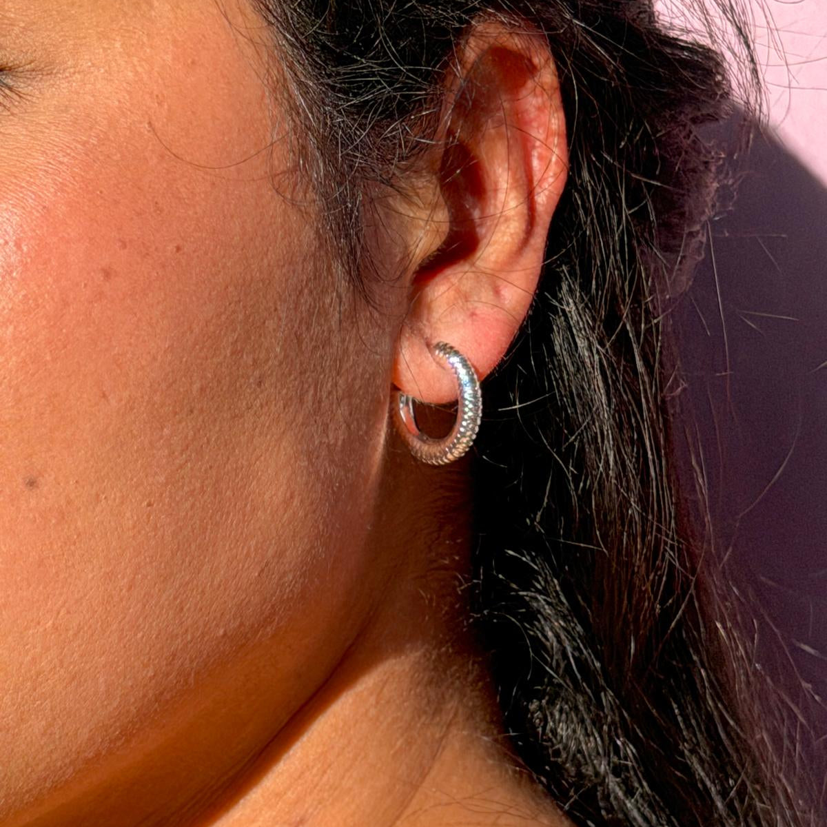 Close-up of an ear wearing a silver hoop earring with a pink background
